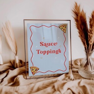 May include: A framed sign with the words "Sauce and Toppings" in red script on a light blue background. The sign is decorated with a red wavy border and cartoon pizza slice illustrations. The frame is a thin, clear metal.