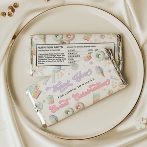 Puede incluir: Un envoltorio de chocolate blanco con un diseño colorido de donuts, conos de helado y caramelos. El envoltorio tiene el texto "Gracias" y "Celebración dulce" en letras rosas y moradas. El envoltorio también incluye una etiqueta de información nutricional con el texto "Amor", "Familia", "Amigos", "Diversión" y "Risa" con un valor diario porcentual del 100%.