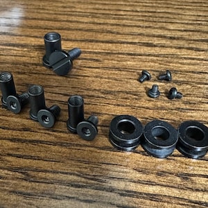 Puede incluir: Un juego de tornillos y arandelas de metal negro. Los tornillos tienen cabeza plana y un accionamiento en forma de estrella. Las arandelas son redondas y tienen un borde ligeramente elevado.