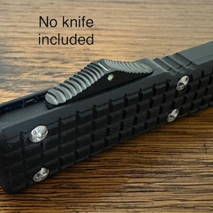 May include: Black tactical-style tool with a textured grip and silver accents. The tool features a sliding mechanism and is labeled "No knife included". The design suggests a focus on functionality and durability.