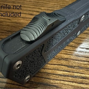 Puede incluir: Mango de cuchillo táctico negro con agarre texturizado y un interruptor de seguridad negro. "Cuchillo no incluido" está escrito en la parte superior de la imagen.