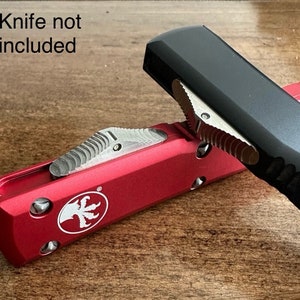 Puede incluir: Dos cuchillos, uno rojo y otro negro, se muestran sobre una superficie de madera. El cuchillo rojo tiene un logotipo con un diseño de pájaro. El texto "Knife not included" es visible en la imagen.