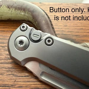 May include: Close-up of a silver folding knife with a partially visible blade. The knife has a matte finish and visible screws. The text "Button only. Knife is not included" is in the image.