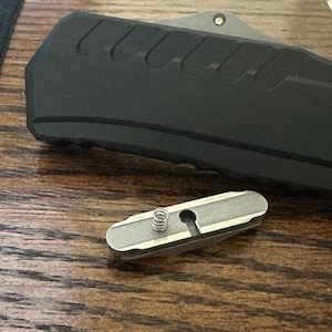 May include: A close-up of a silver metal spring and a metal plate with a hole in the center. The plate is on a wooden surface.