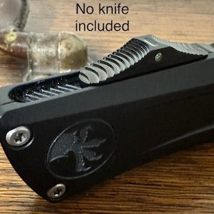 Puede incluir: Funda de cuchillo táctico negra con una superficie texturizada y un pequeño logotipo plateado. La funda tiene una mira elevada en la parte superior. El texto "No knife included" es visible en la imagen.