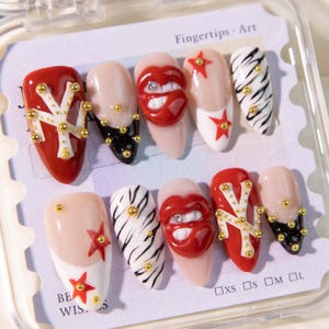 May include: A set of press-on nails with various designs. Some nails are red with gold accents, others feature a lip design with rhinestones, and some have a zebra stripe pattern. The text "Fingertips Art" is visible.