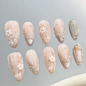 May include: A set of ten artificial nails in a soft pink hue, adorned with delicate white floral embellishments, gold accents, and shimmering glitter. The nails are oval-shaped, offering a sophisticated and feminine aesthetic.