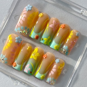 May include: A set of ten false nails with a floral design. The nails are painted in a light peach color with a gradient of yellow and blue. The flowers are painted in shades of yellow, blue, and pink. The nails are decorated with small rhinestones.