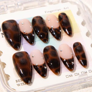 May include: A set of artificial nails with a tortoiseshell design and a nude base. Each nail is adorned with small, silver rhinestones. The nails are packaged in a clear plastic container with the text "Fingertips - Art" and "Best Wishes".
