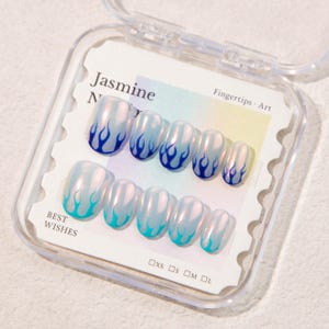 Puede incluir: Un juego de uñas postizas ovaladas con un diseño de llama azul. Las uñas están en una caja de plástico transparente con el texto "Jasmine N" y "Best Wishes". El diseño de la llama pasa de azul claro a azul oscuro.