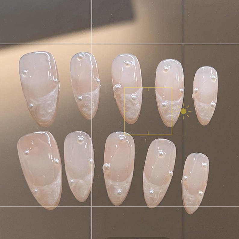 May include: Ten nude-colored oval-shaped fake nails with white pearl accents.