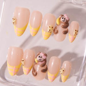 May include: A set of artificial almond-shaped nails. Each nail has a light pink base with a yellow and pink checkered design at the tip. Some nails feature a 3D bear charm. The bears are brown and tan with pink bows.