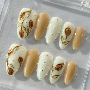 May include: Set of ten false nails with a floral design. The nails are in two different styles: nude with gold accents and white with gold accents. The nails are decorated with small gold flowers and gold dots.