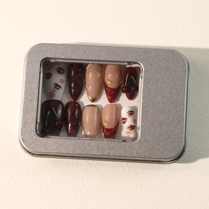 May include: A set of ten press-on nails in a silver rectangular metal tin. The nails feature various designs including white with red lip prints and gold studs, dark red leopard print, solid nude with gold tips, and solid dark red with gold star accents.