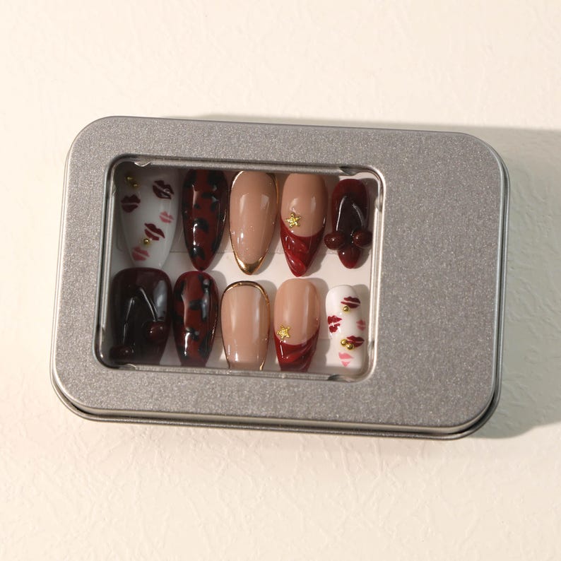 May include: A set of ten press-on nails in a silver rectangular metal tin. The nails feature various designs including white with red lip prints and gold studs, dark red leopard print, solid nude with gold tips, and solid dark red with gold star accents.