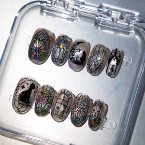 May include: A set of ten artificial nails with various designs. The nails feature stained glass, cat silhouettes, and floral patterns in a clear, iridescent finish. The nails are displayed in a clear plastic container.