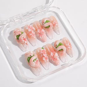 Puede incluir: Un juego de diez uñas artificiales en una caja de plástico transparente. Cada uña presenta un diseño degradado rosa y blanco, adornado con adornos de flamencos y flores en 3D. Los flamencos son verdes y rosas, y las flores son rosas con acentos de perlas.