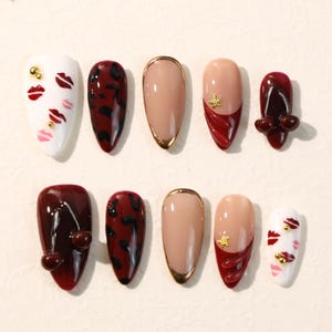 May include: A set of ten press-on nails featuring almond-shaped tips in various designs, including white with red lip prints and gold studs, dark red leopard print, nude with gold trim, nude with red tips and gold stars, and dark red with 3D accents.