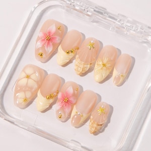 May include: A collection of ten artificial nails presented in a clear case. The nails are primarily light pink, adorned with designs such as pink and white flowers, gold beads, and seashell patterns. Some nails have a pale yellow tip.