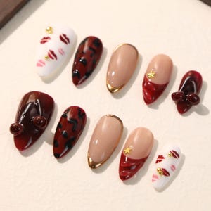 May include: A set of ten press-on nails featuring various designs including white nails with red lip prints and gold studs, dark red leopard print nails, nude nails with gold tips, and dark red nails with 3D cherry or star embellishments.
