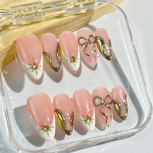May include: A set of ten almond-shaped artificial nails. Each nail features a pale pink base with a white tip and gold accents. Some nails have a gold bow design, while others have a gold starburst.
