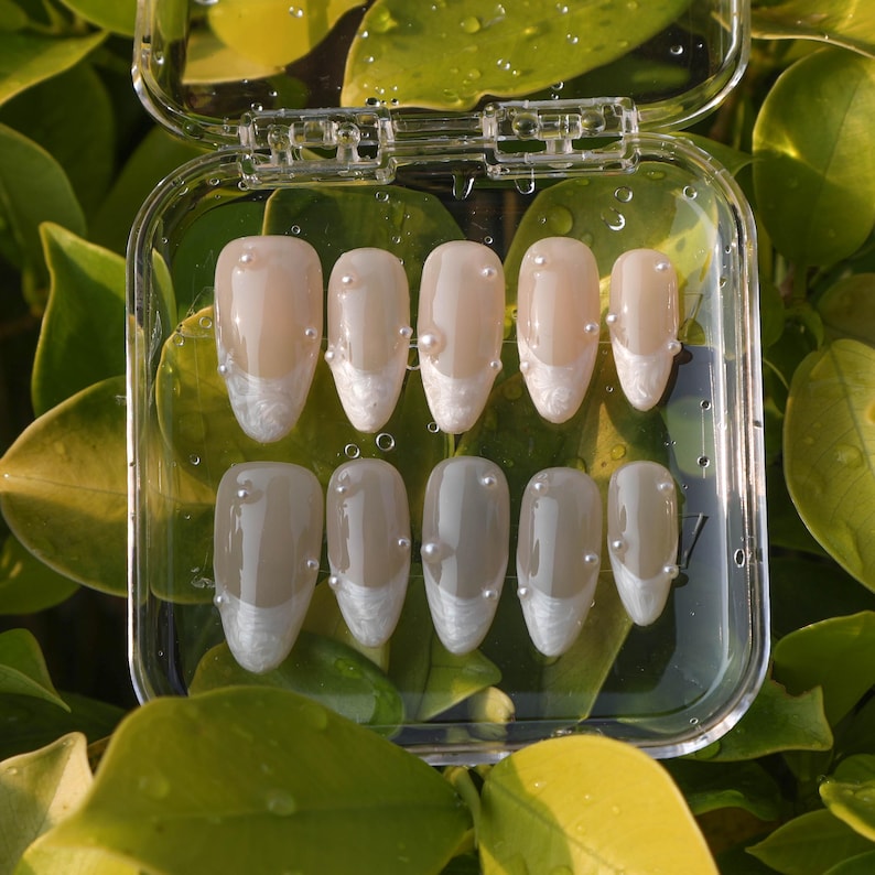 May include: A set of ten false nails in a clear plastic container. The nails are oval shaped and have a white tip with a pearl accent. The nails are arranged in two rows of five.