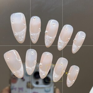May include: Ten nude-colored fake nails with a white design and pearl accents.