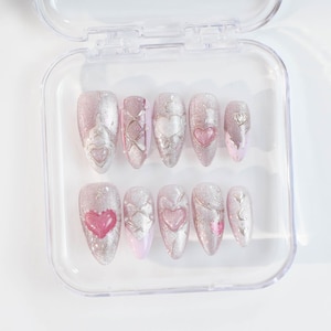 May include: A set of ten pink and silver glitter press-on nails with heart designs. The nails are arranged in a clear plastic case.