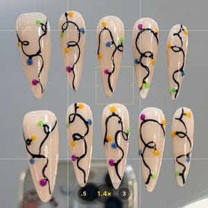 Peut inclure: Dix faux ongles de couleur nude avec un motif de guirlandes lumineuses noires avec des ampoules colorées. Les lumières sont disposées en un motif incurvé sur chaque ongle.