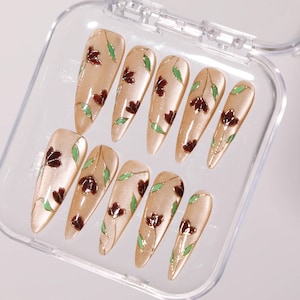 May include: A set of ten almond-shaped false nails in a clear plastic case. Each nail is a light beige with a floral design featuring burgundy flowers, green leaves, and gold accents. The nails are arranged in a neat row.