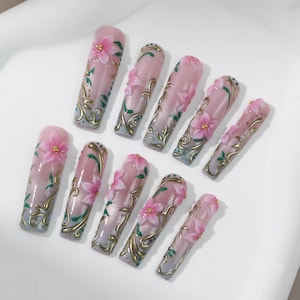 May include: A set of ten artificial nails with a pink and white gradient design. Each nail features a 3D pink flower, gold filigree, and green leaf accents. The nails are arranged on a white surface.