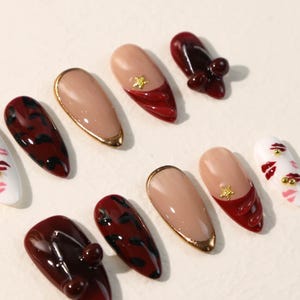 May include: A set of ten almond-shaped press-on nails featuring various designs including deep red cherry motifs, dark brown leopard print, nude bases with gold metallic tips, and white nails decorated with small red lip prints and gold studs.
