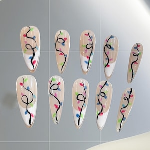 Peut inclure: Dix faux ongles de couleur nude avec une pointe blanche et un motif de lumières de Noël colorées sur chaque ongle.