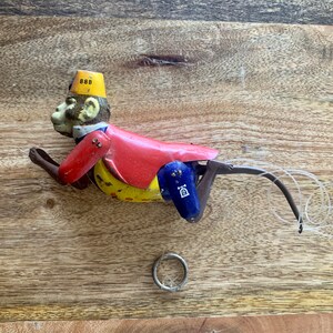 May include: A vintage tin toy monkey wearing a yellow and red hat with the number 888 on it. The monkey is painted in red, yellow, blue, and black. The monkey is in a crouching position with its arms outstretched and its tail curled up. The toy is missing a piece of its tail.