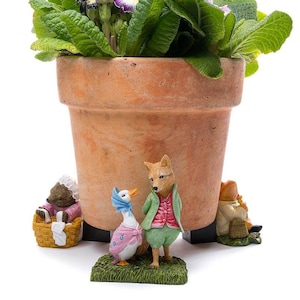 May include: A terracotta flower pot with green leafy plants and purple and white flowers. The pot has three small figurines at the base, including a fox, a duck, and a rabbit.