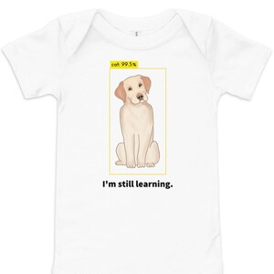 Könnte beinhalten: Weißer Baby-Strampler mit einem gelben Rahmen um ein Cartoon-Bild eines Golden Retriever-Hundes. Der Text "cat: 99.5%" befindet sich in der oberen linken Ecke des Rahmens. Der Text "I'm still learning." befindet sich unter dem Rahmen.
