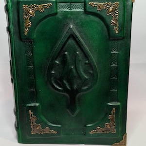 May include: A dark green leather-bound book featuring an embossed spade-shaped emblem in the center. The cover is decorated with intricate geometric patterns and four ornate brass-colored metal corner protectors with filigree detailing.