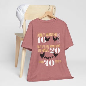 Op de afbeelding: Een stoffig roze t-shirt met de tekst "I ONLY WANTED 10 CHICKENS BUT IF FATE WANTS ME TO HAVE 20 THEN 40 IT IS!!!" in wit en geel. Het shirt heeft zwarte kippensilhouetten.