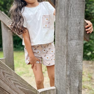 Könnte beinhalten: Ein weißes T-Shirt mit kurzen Ärmeln und einer Tasche mit Gänseblümchen-Druck sowie passende beige Shorts mit Gänseblümchen-Druck.