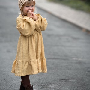 May include: A young girl wearing a beige crocheted hat and a yellow dress with a ruffled hem. She is standing on a paved road and smiling.