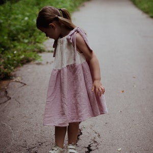 Puede incluir: Una niña pequeña con un vestido rosa y blanco con un dobladillo con volantes y zapatillas blancas de pie en un camino de asfalto agrietado.