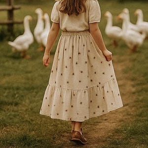 Könnte beinhalten: Eine junge Person trägt einen Strohhut und ein weißes Kleid mit braunem Sternenmuster und geht von einer Gruppe weißer Gänse weg. Das Kleid hat einen Stufenrock und die Person trägt braune Schuhe.