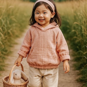 May include: A young girl wearing a pink hooded sweatshirt and beige pants walks down a path carrying a wicker basket with a plush bunny inside. She is wearing a pink headband and brown shoes.