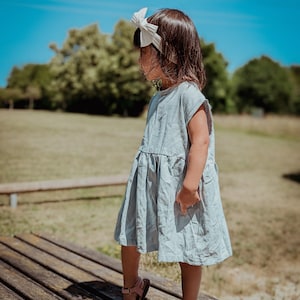 Puede incluir: Una niña pequeña con un vestido azul claro y una diadema blanca con un lazo camina sobre una plataforma de madera. Lleva sandalias rosas con adornos de flores.