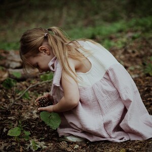 May include: A young girl in a pink and white dress is kneeling in a forest setting, looking down at the ground. She is holding a small cluster of pine cones in her hand.
