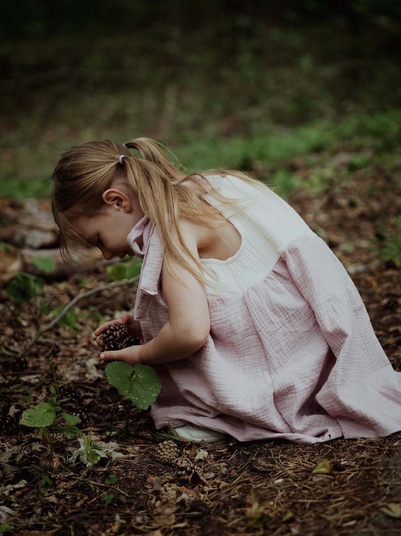 May include: A young girl in a pink and white dress is kneeling in a forest setting, looking down at the ground. She is holding a small cluster of pine cones in her hand.