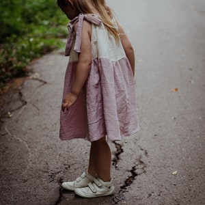 May include: A young girl wearing a pink and white dress with a ruffled skirt and white sneakers stands on a cracked asphalt path.