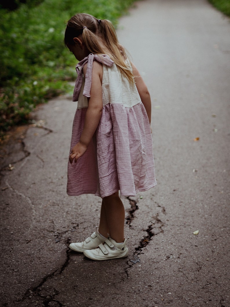May include: A young girl wearing a pink and white dress with a ruffled skirt and white sneakers stands on a cracked asphalt path.