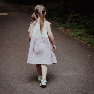 May include: A young girl in a pink and white dress walks down a paved path in a forest. She has her hair pulled back in pigtails and is wearing white shoes.