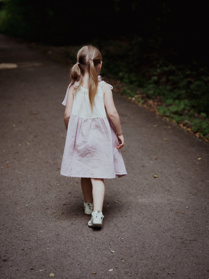 May include: A young girl in a pink and white dress walks down a paved path in a forest. She has her hair pulled back in pigtails and is wearing white shoes.